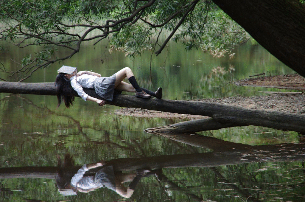 A woman sleeping idly on a tree trunk leaning over water.